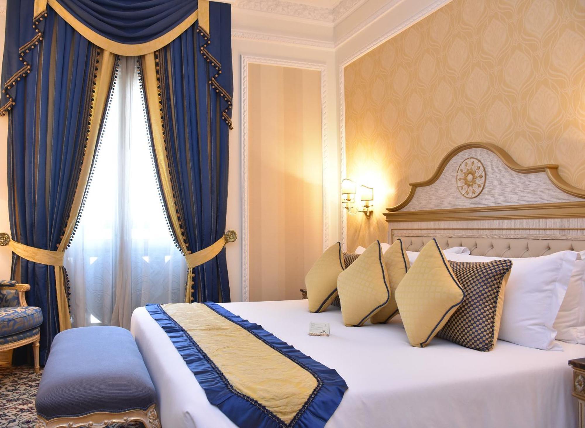 Royal Rose Abu Dhabi, A Curio Collection By Hilton Affiliated Hotel Exterior photo The photo shows a luxurious hotel room. It features a large bed with multiple decorative pillows in shades of gold and blue. There is a decorative bed runner that matches these colors. To the side of the bed, there is a small upholstered bench. The c